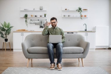 Rahat bir kanepede otururken kablosuz bilgisayarda yazan günlük kıyafetlerle odaklanmış beyaz bir adam. Gözlüklü çekici erkek, evde otururken internette çalışıyor..