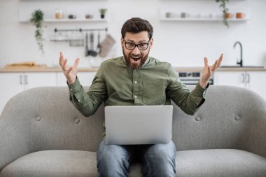 Şaşkın sakallı erkek, gözlük takıyor ve gündelik giysiler giyiyor, omuzlarını silkiyor ve modern dizüstü bilgisayarla kanepeye oturuyor ve kameraya bakıyor. Şüphe kavramı, insanlar ve teknoloji.