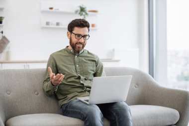 Kanepede oturan yakışıklı serbest çalışan ve evdeki video konferansı için modern dizüstü bilgisayar kullanan. Gözlüklü kendine güvenen sakallı adam web kamerasından konuşuyor ve elleriyle hareket ediyor..