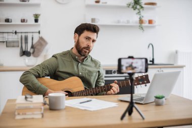 Masada oturup tripodla modern akıllı telefondan video kaydederken gitar çalan beyaz adam virtüözünden esinlendim. Erkek vlogger sosyal medyaya enstrüman teknikleri hakkında yeni bir ders oluşturuyor.