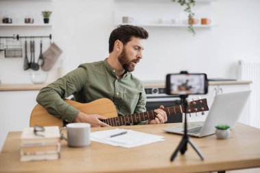Masada oturup tripodla modern akıllı telefondan video kaydederken gitar çalan beyaz adam virtüözünden esinlendim. Erkek vlogger sosyal medyaya enstrüman teknikleri hakkında yeni bir ders oluşturuyor.