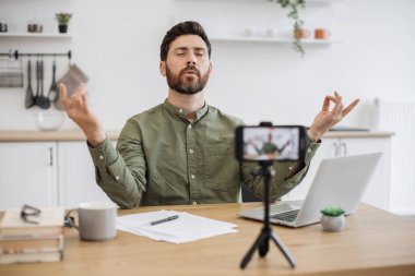 Akıllı telefonun önünde oturan konsantre sakallı vlogger tripoda sabitlenmiş ve evde canlı yayın yapıyor. Sakin adam Mudra jestinde el ele tutuşuyor ve yoga meditasyonu dersi veriyor..