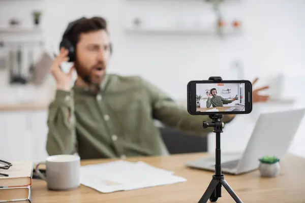 Attractive bearded vlogger listening podcasts and keeping hand in wireless headphones on blurred background. Focus on modern cell phone fixed on tripod with active screen.