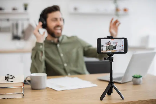 Modern smartphone fixed on tripod recording satisfied blogger wearing wireless headphones and dancing with hands. Confident content maker sitting in front of laptop and enjoying creative process.