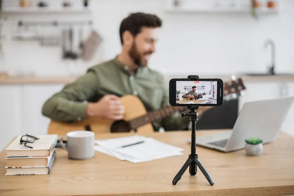 Skillful musician sitting at home office using smartphone and tripod for filming video tutorial about guitar playing. Handsome male blogger sharing with followers in social media artistic experience.