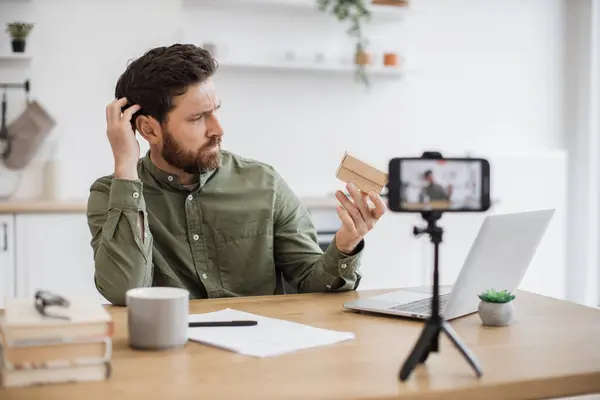 Caucasian bearded man scratching head while looking with curiosity on cardboard box. Handsome male blogger making unpacking of online order while recording video on smartphone.