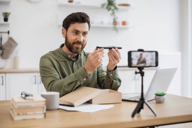 Heyecanlı blogcu, web mağazasından yeni akıllı telefon alımıyla birlikte posta paketini açarken masada oturuyor. Mutlu genç erkek paket teslimatı yapıyor ve cep telefonuna video kaydediyor..