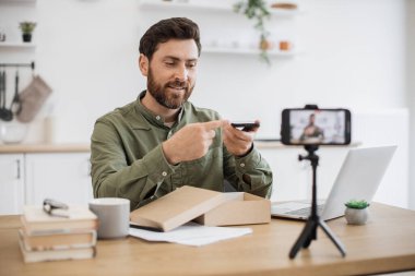 Heyecanlı blogcu, web mağazasından yeni akıllı telefon alımıyla birlikte posta paketini açarken masada oturuyor. Mutlu genç erkek paket teslimatı yapıyor ve cep telefonuna video kaydediyor..