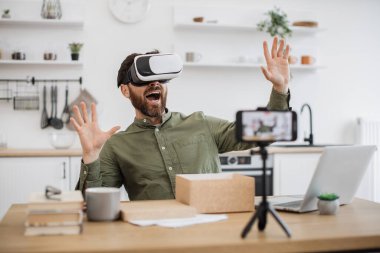 Evde yeni VR kulaklığı incelerken modern akıllı telefonu tripodla kullanan yakışıklı bir teknoloji blogcusu. Beyaz tenli erkek etkili boks aleti açıyor ve sosyal medya için içerik yaratıyor.