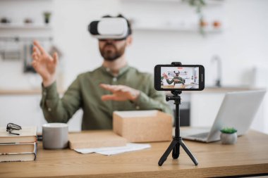 Kafkas erkek vlogger bulanık arka planı VR kulaklık ile masada oturuyor ve canlı yayın yapıyor. Tripoda sabitlenmiş modern akıllı telefona odaklan. Satın alma, teknoloji ve sosyal medya kavramı.