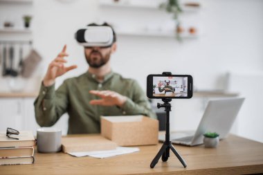 Kafkas erkek vlogger bulanık arka planı VR kulaklık ile masada oturuyor ve canlı yayın yapıyor. Tripoda sabitlenmiş modern akıllı telefona odaklan. Satın alma, teknoloji ve sosyal medya kavramı.