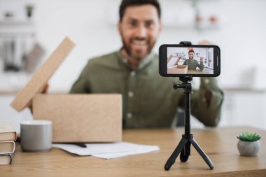 Evde karton kutuları açarak enerjik sakallı adamı kaydeden akıllı telefon ekranına odaklan. Kafkas erkek vlogger paylaşımı sosyal medyada abonelerle yeni online satın alma.