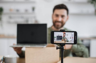 Tripoda sabitlenmiş modern akıllı telefon pozitif sakallı adamın yeni alınmış dizüstü bilgisayarının paketini açtığı videoyu kaydediyor. Gülümseyen Kafkasyalı vlogger modern aygıtlar hakkında geri bildirim ve tavsiyelerde bulunuyor.