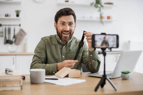 Caucasian male influencer feeling excitement and happiness while advertising his lovely purchase on smartphone camera. Amazed confident man comparing prices of unisex watches during video recording.