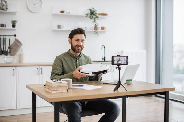 Caucasian blogger recording video content on modern smartphone while unboxing new robot vacuum cleaner at home. Smiling influencer conducting review on modern gadget in front of mobile camera.