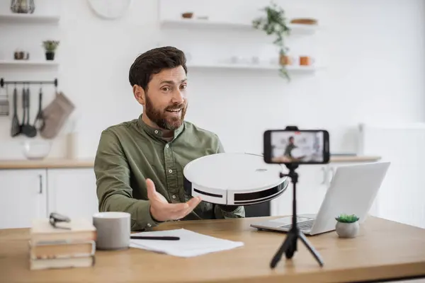 stock image Caucasian blogger recording video content on modern smartphone while unboxing new robot vacuum cleaner at home. Smiling influencer conducting review on modern gadget in front of mobile camera.