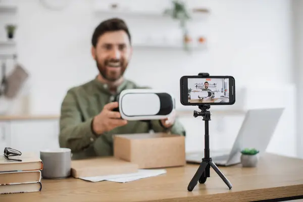 Happy caucasian man showing virtual reality glasses while filming himself of modern smartphone standing on tripod. Handsome technology blogger working remotely in personal cabinet at home.