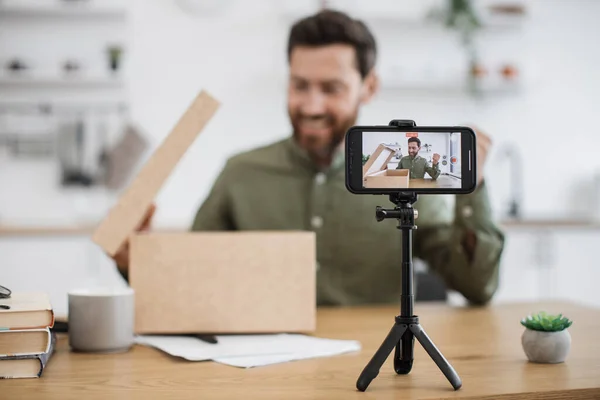 Focus on smartphone screen recording energetic bearded man doing unpacking of cardboard box at home. Caucasian male vlogger sharing with subscribers in social media new online purchase.