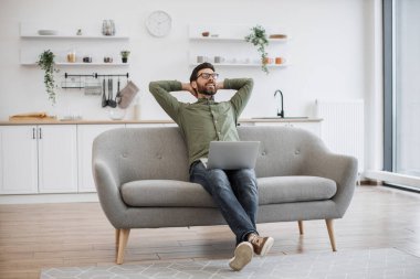 Pozitif adam ellerini başının arkasına koyup rahat bir kanepeye uzanıp tatilin hayalini kuruyor. Gözlüklü ve günlük kıyafetli beyaz sakallı bir erkek evde otururken uzaktan çalışmanın tadını çıkarıyor..