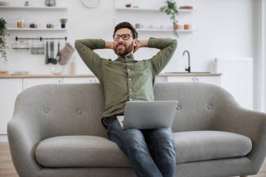 Gündelik giysiler ve gözlüklü beyaz sakallı bir adam rahat koltukta oturuyor ve başının arkasında el ele tutuşuyor. Erkek serbest çalışan rahat evdeki online işten sonra dinleniyor..