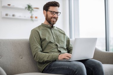 Rahat bir kanepede otururken kablosuz bilgisayarda yazan günlük kıyafetlerle odaklanmış beyaz bir adam. Gözlüklü çekici erkek, evde otururken internette çalışıyor..