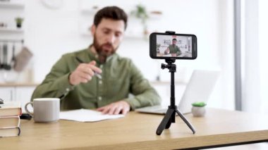 Sosyal medyada yeni video içeriği yaratmak için akıllı telefon ve tripodu kullanan mutlu sakallı adam. Günlük giysiler içindeki olumlu erkek blogcu çevrimiçi iletişim sırasında abonelerle etkileşime giriyor.