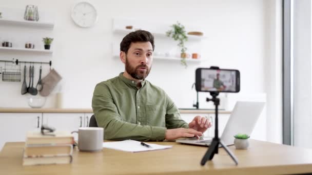 現代のスマートフォンと三脚を使用したアクティブな男性インフルエンサー ホームオフィスからのライブストリーミング デスクに座っているカーキシャツのハンサムなひげ付き男 ラップトップを入力してモバイルWebカメラで話す — ストック動画