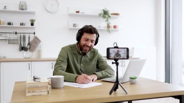 キッチンに座って ノートを作っている間スマートフォンカメラを見ているヘッドフォンを持つポジティブなブルネット男 オリーブシャツの楽観的な慎重なブロガー 笑顔とビデオを録画する — ストック動画