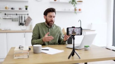 Yakışıklı sakallı blogcu mutfak masasında oturmuş kameralı akıllı telefondan video çekerken cihazlar kullanıyor. Kafkasyalı erkek heyecanlı bir şekilde elindeki küçük kutuya bakıyor..
