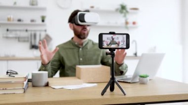 Mutlu erkek blog yazarının yeni VR kulaklığını test ettiği ve sosyal medyadaki aboneler için inceleme yaptığı modern akıllı telefon kaydı videosu. Yenilikçi elektronik ve çevrimiçi satın alma kavramı.