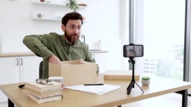 Gündelik kıyafet giyen mutlu erkek blogcu yeni kablosuz dizüstü bilgisayarı olan bir kutu açıyor ve tripodlu modern akıllı telefon kullanarak video kaydediyor. Çevrimiçi alışveriş, sosyal medya ve teknoloji kavramı.
