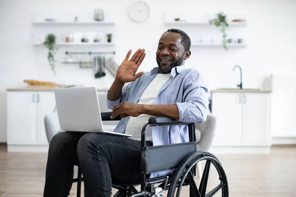 Cheerful adult waving hello at webcam of portable computer while greeting colleague in home office. Young african person in casual wear sitting in wheelchair in studio apartment.