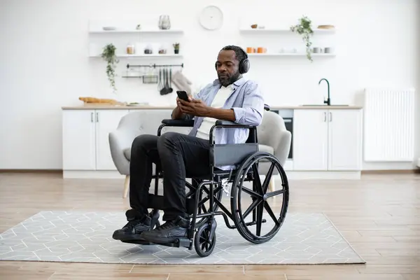 Relaxed african person in headphones holding smartphone while sitting in wheelchair in studio apartment. Calm homeowner checking playlist before listening to music over digital devices at home.