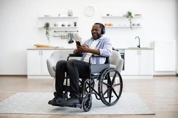 Relaxed african person in headphones holding smartphone while sitting in wheelchair in studio apartment. Calm homeowner checking playlist before listening to music over digital devices at home.