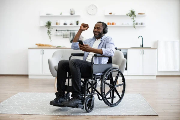 Joyous man in button-down shirt raising hand while looking at smartphone sitting in wheelchair in studio apartment. Cheerful african adult dancing along while hearing loud tunes in headphones.