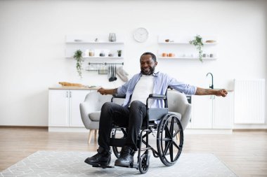 Tekerlekli sandalyedeki neşeli sakallı adam evde egzersiz yaparken kollarını esnetiyor. Konforlu giysiler içinde Afro-Afrikalı bir yetişkin dairede fiziksel aktivitelere katılıyor..
