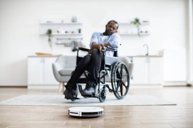 Kapalı alanda otomatik temizlik yaparken hareketlilik bozukluğu olan mutlu Afrikalı adam tarafından işletilen akıllı robotik vakuma odaklanın. Neşeli ev sahibi uzaktan kumandayla ev aletini programlıyor.