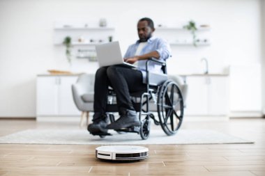 Seçici odak noktası robotik vakum. Halıdaki tozu temizlerken Afrikalı ev sahibi dizüstü bilgisayar kullanıyor. Tekerlekli sandalyedeki serbest çalışan, teknolojik aletler kullanarak rahat bir hayat sürüyor.