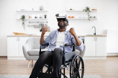 Güneşli bir günde evde keyifli bir şekilde otururken tekerlekli sandalyeli, VR kulaklıklı Afrikalı bir erkek. Özürlü gülümseyen olgun bir insan VR kulaklık takıyor ve el kol hareketi yapıyor..