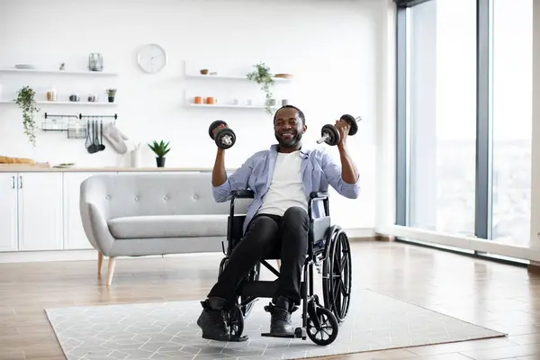 Full length view of handsome african man in wheelchair lifting weights while exercising with limited mobility indoors. Smiling adult using moderate-intensity activity during home workout in kitchen.