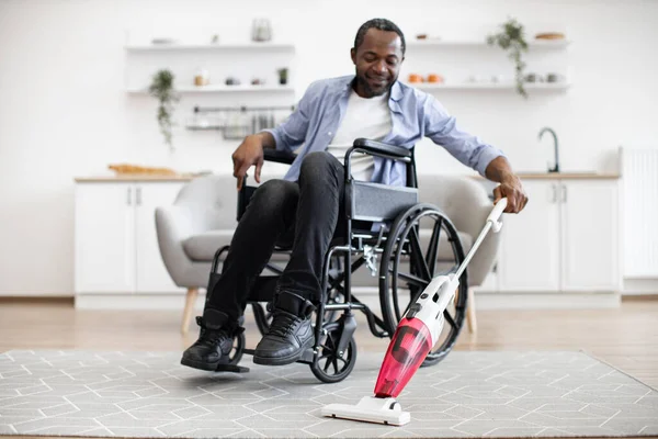 Smiling man with mobility impairment handling debris on carpet with wireless vertical vacuum cleaner. Joyful african adult in casual clothes having fun with daily housekeeping routine.