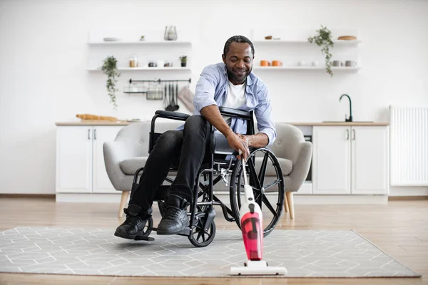 Smiling man with mobility impairment handling debris on carpet with wireless vertical vacuum cleaner. Joyful african adult in casual clothes having fun with daily housekeeping routine.
