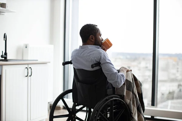 Back view of african american wheelchair user with cup of hot drink looking at panoramic window resting in open-plan kitchen of modern apartment.