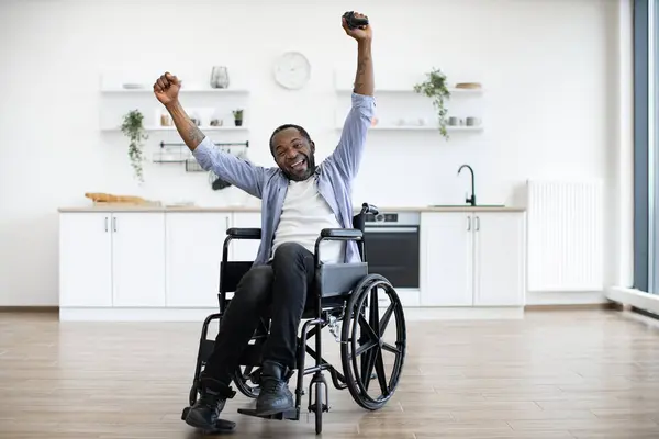 Full length of african wheelchair user celebrating success by raising hands with joystick in air. Cheerful young man playing video games for competition and computer learning.