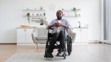 Modern dairenin açık planlı mutfağında dinlenirken kameraya gülümseyen Afro-Amerikan tekerlekli sandalye kullanıcısının tam boy portresi. Günlük giysiler içinde neşeli genç erkek içeride zaman geçiriyor..