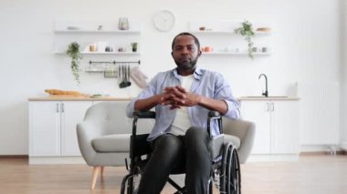Modern dairenin açık planlı mutfağında dinlenirken kameraya gülümseyen Afro-Amerikan tekerlekli sandalye kullanıcısının portresini yakından çek. Neşeli genç erkek, günlük giysiler içinde zaman harcıyor, başparmağını kaldır.