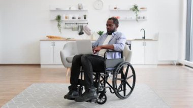 Tekerlekli sandalyedeki neşeli Afrikalı adam dizüstü bilgisayarını tutarken açık-plan mutfağın ortasında kalıyor. Evdeki kazadan sonra iyileşme sürecinde internet sayfalarında mutlu genç sörfçüler..