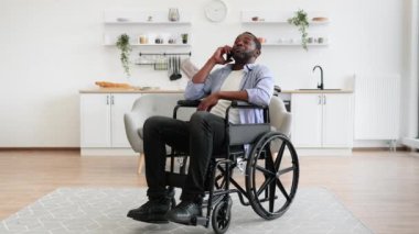 Modern stüdyo dairesinin rahatlığını kullanırken tekerlekli sandalyedeki yetişkin birinin cep telefonuyla konuşmasının geniş açılı görüntüsü. Olgun Afrikalı adam hastalıktan kurtulurken doğum günü partisi düzenliyor..