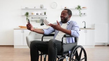 Modern apartmanda gezmeyi planlarken engelli cep telefonlu neşeli genç bir yetişkin. Gülümseyen Afrikalı bir adam yemek odasında video başvurusu yoluyla akrabalarıyla sohbet ediyor..