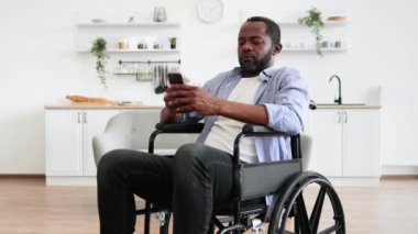 Sıradan giyimli, akıllı telefonlu, tekerlekli sandalyede stüdyo dairesinde oturan genç bir Afrikalı Amerikalı. Olumlu yetişkin bir adam harika bir fikirden esinlenerek çenesine dokunur..